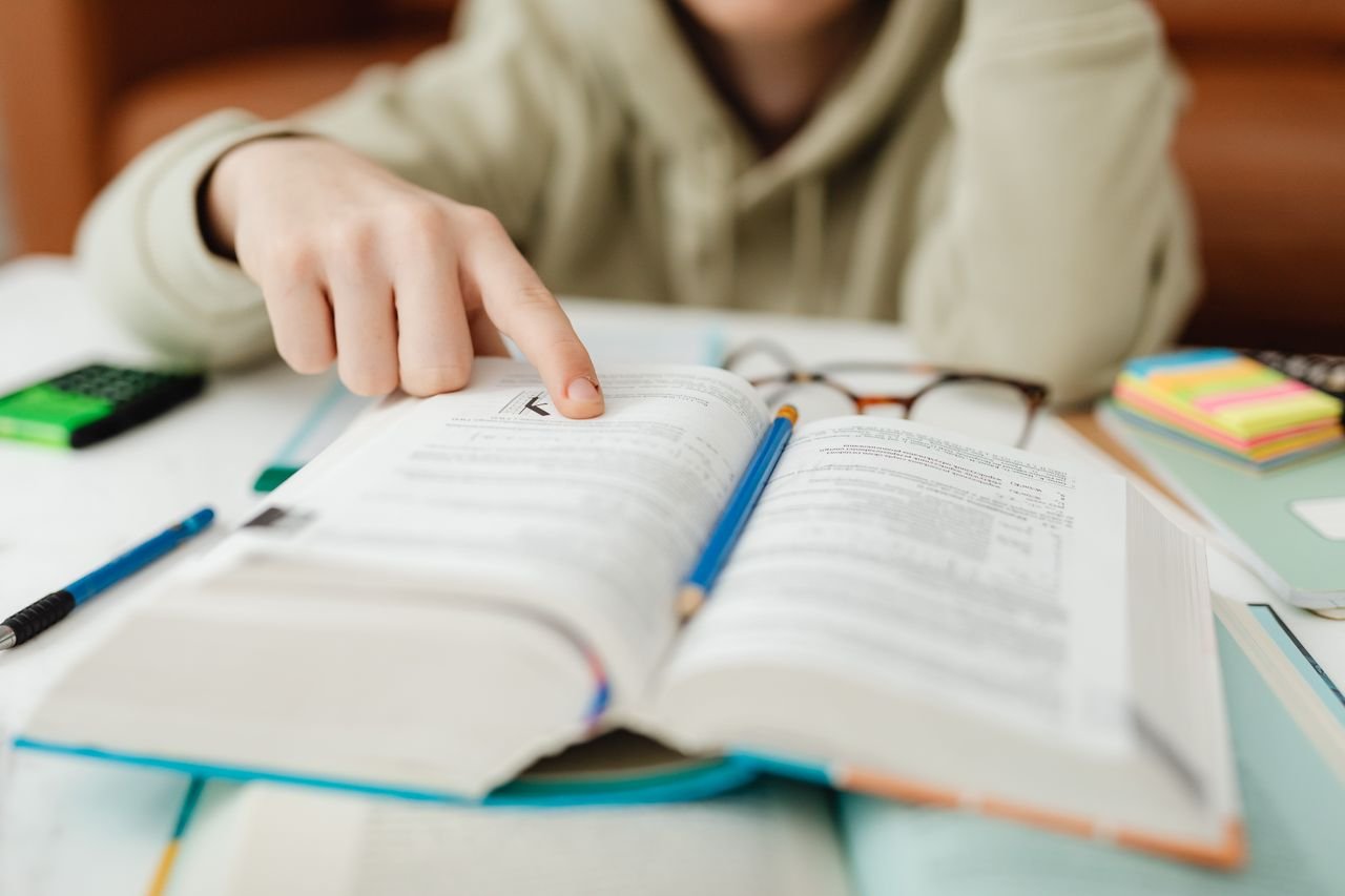 Focused student pointing at text in an open book, studying for exams indoors.